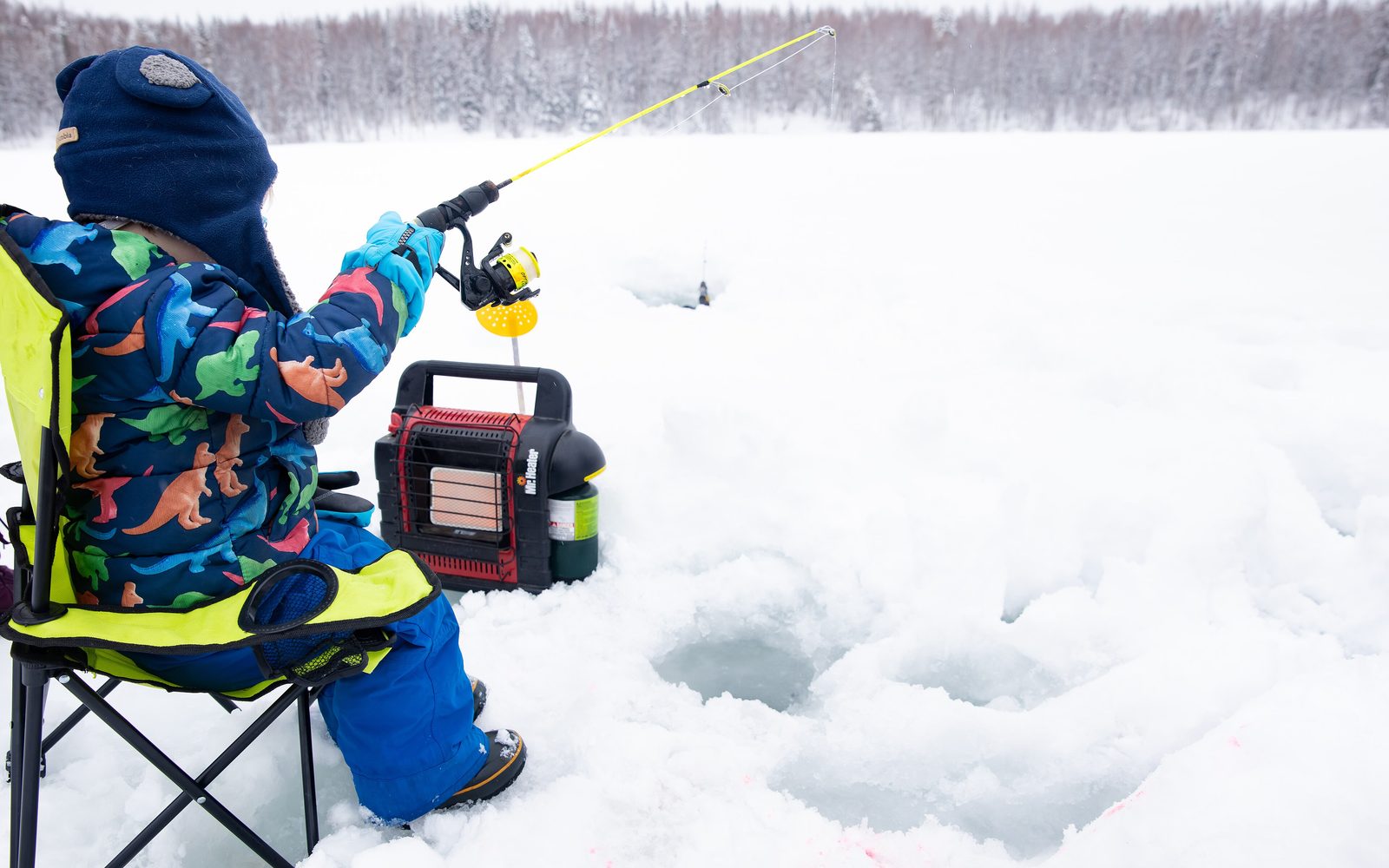 Child in a snowsuit jigging a short rod over a hole in the ice, with a portable heater on the snow beside the camp chair