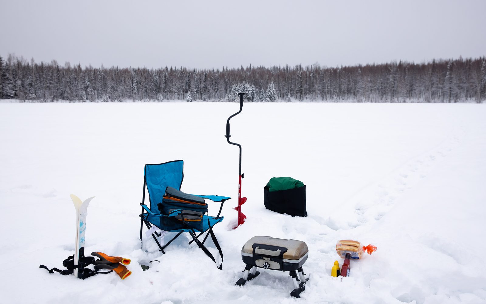 Ice fishing gear set up on a snow-covered frozen lake: a hand auger, a camp chair and a portable grill