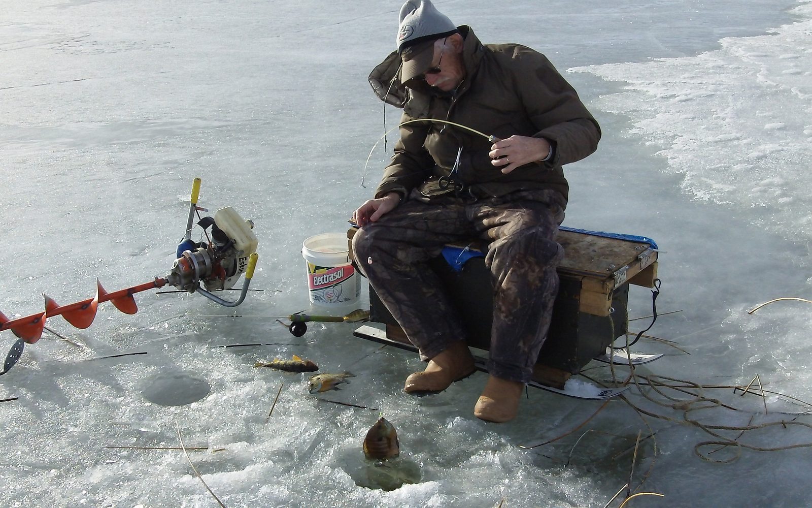 Angler seated on a bucket beside a hand-held power auger and an open hole in the ice
